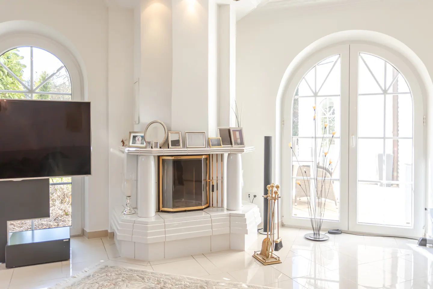 Bright living room with white walls, tile floor, and arched windows. A white fireplace with gold trim is centered, decorated with photos. A TV is on the left.