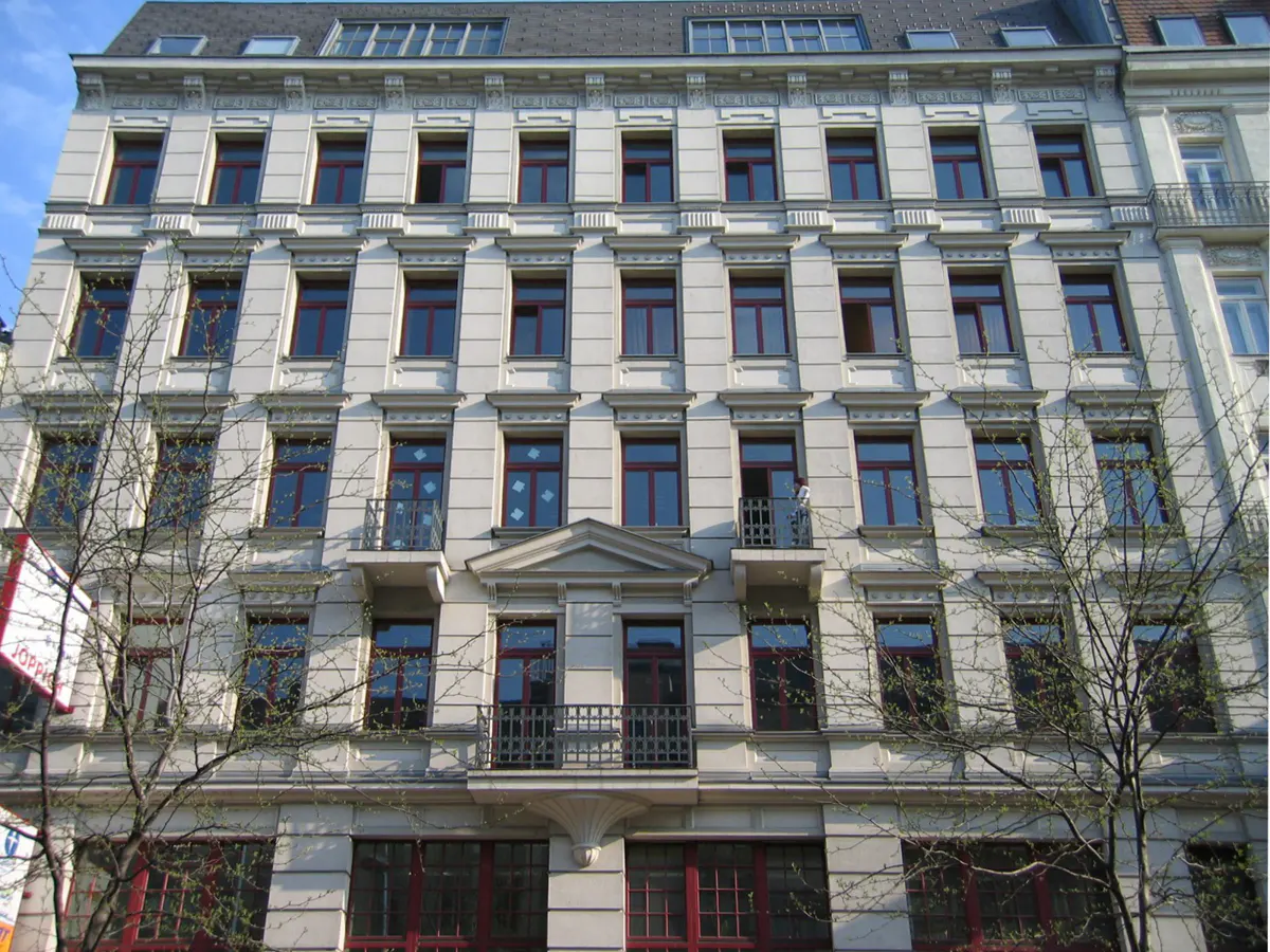 A five-story apartment building with white walls and red window frames. Balconies are on the third and fourth floors.