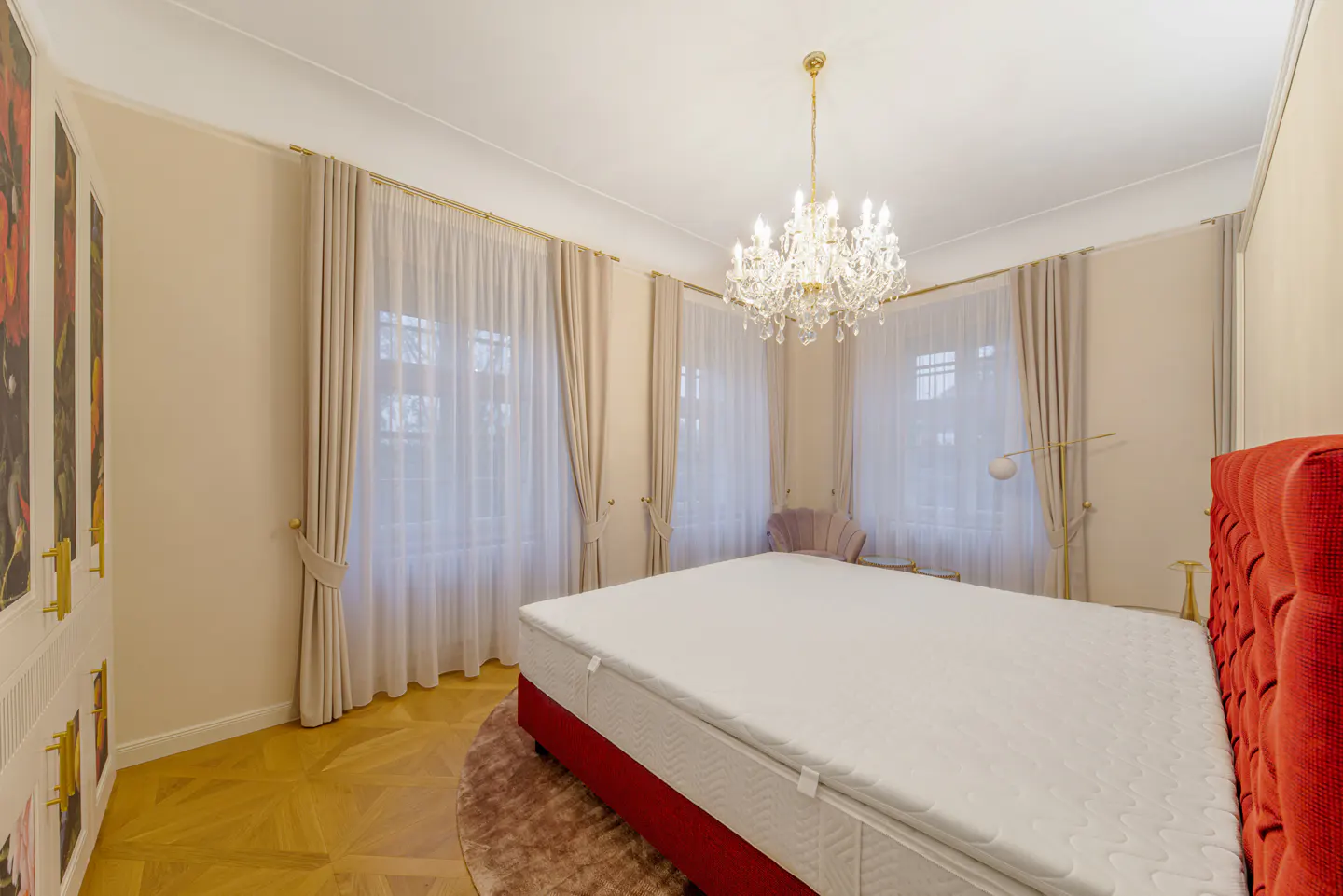 Bright bedroom with a red bed frame, white bedding, and a crystal chandelier. Windows with sheer curtains and a floral wardrobe are also visible.