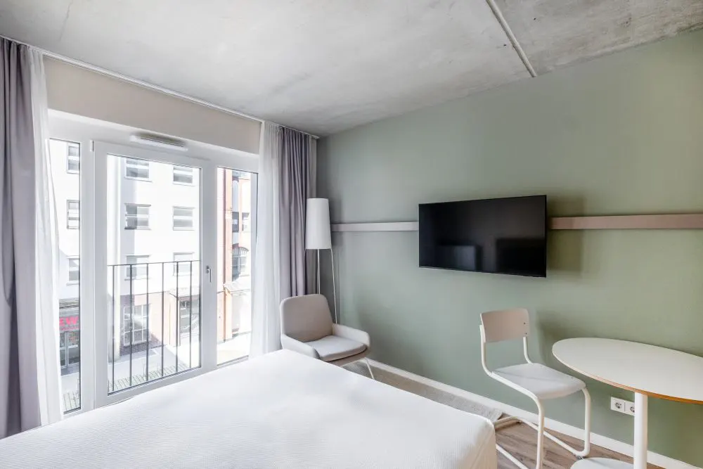 A modern hotel room with a white bed, balcony, chair, table, and a wall-mounted TV on a light green wall.