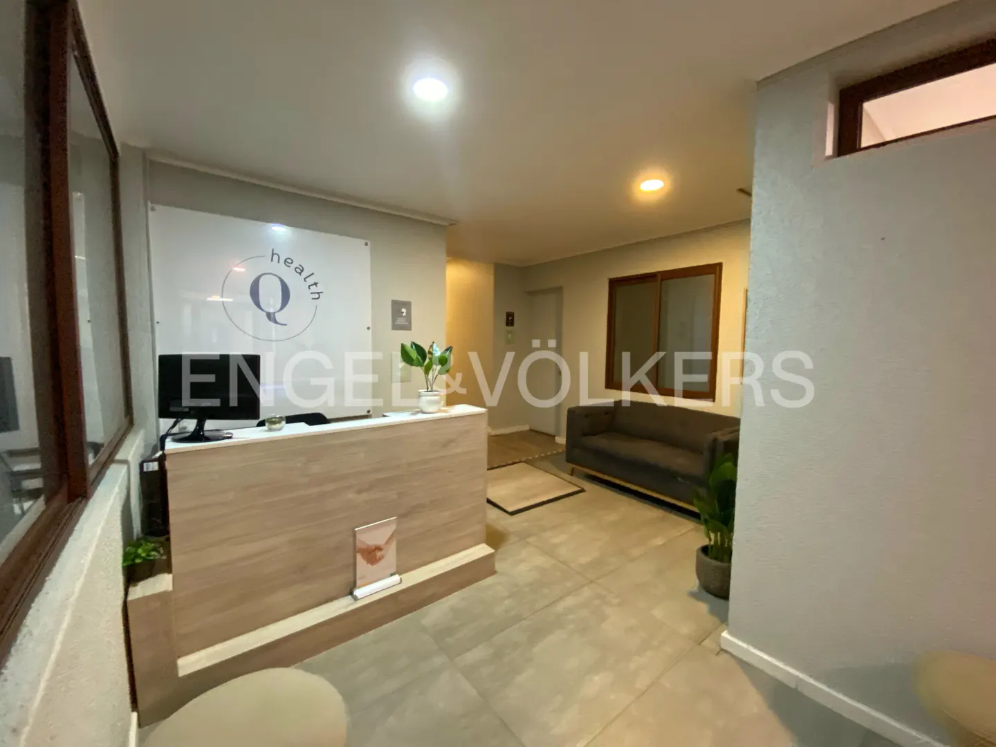 Modern office lobby with a light wood reception desk, computer, and waiting area with a gray sofa.