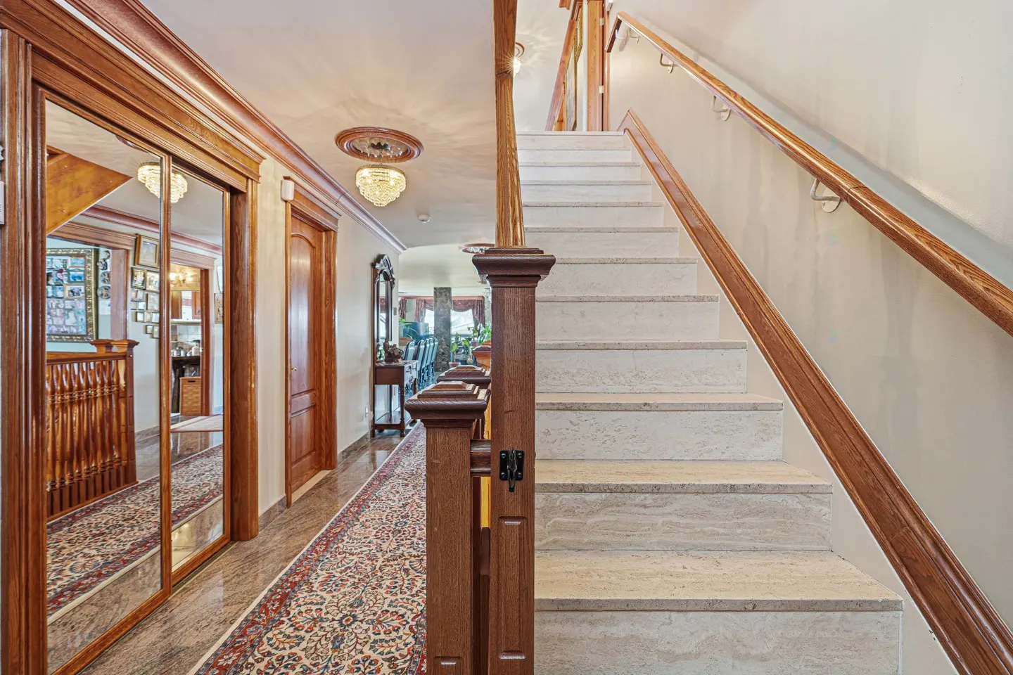 Hallway with marble stairs, wood railing, and patterned rug. Mirrored closet doors reflect a room with framed photos. Chandelier visible.