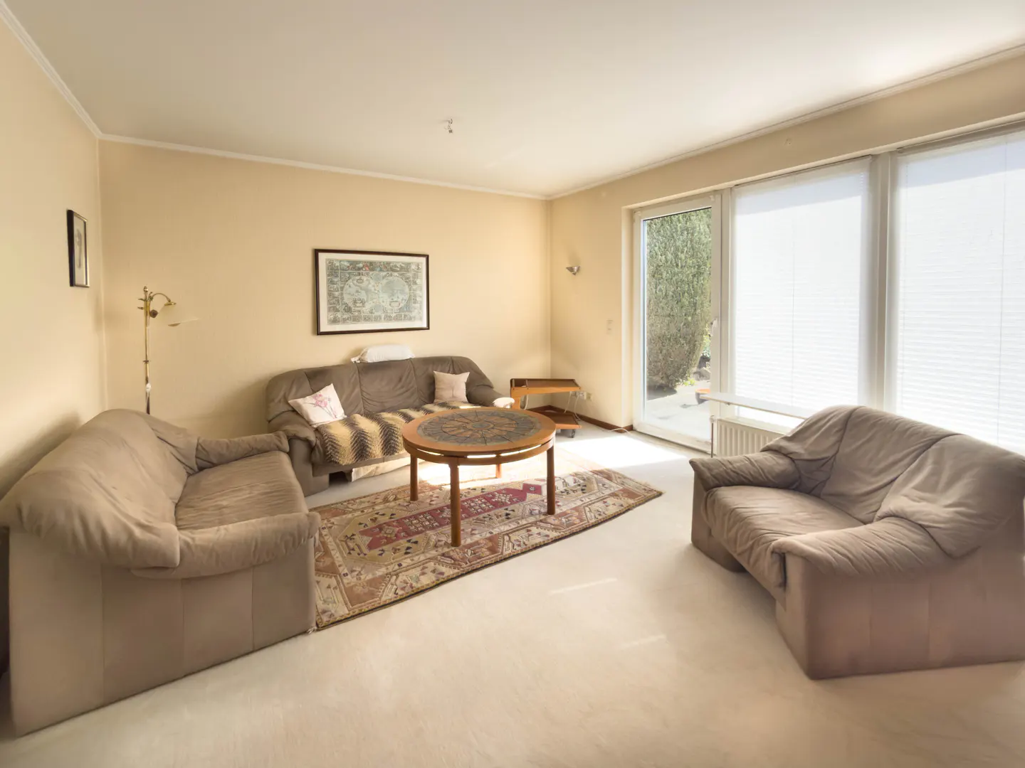 Living room with beige walls, white carpet, brown sofas, and a round wooden table on a patterned rug. Large windows with blinds let in natural light.