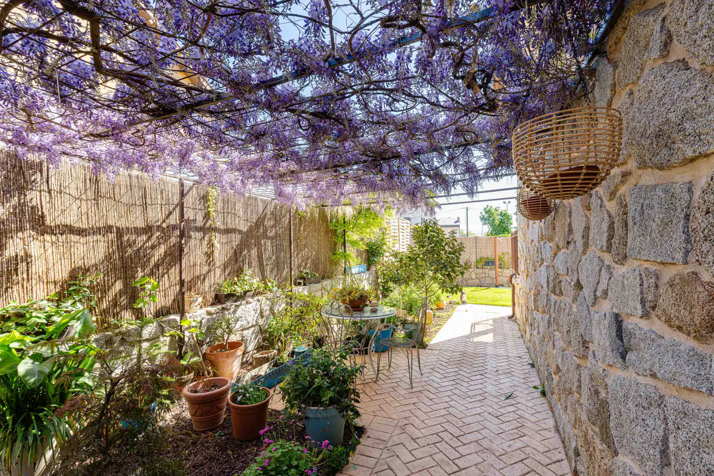 A charming patio with a stone wall, brick floor, and a wisteria-covered pergola. Potted plants and a small table add to the cozy atmosphere.