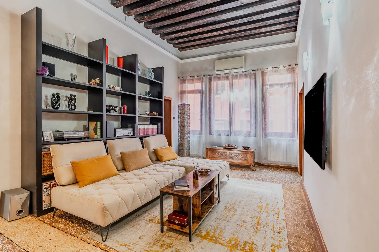 Living room with a beige sofa, wooden ceiling beams, and a black bookshelf filled with books and decor. A TV hangs on the wall.