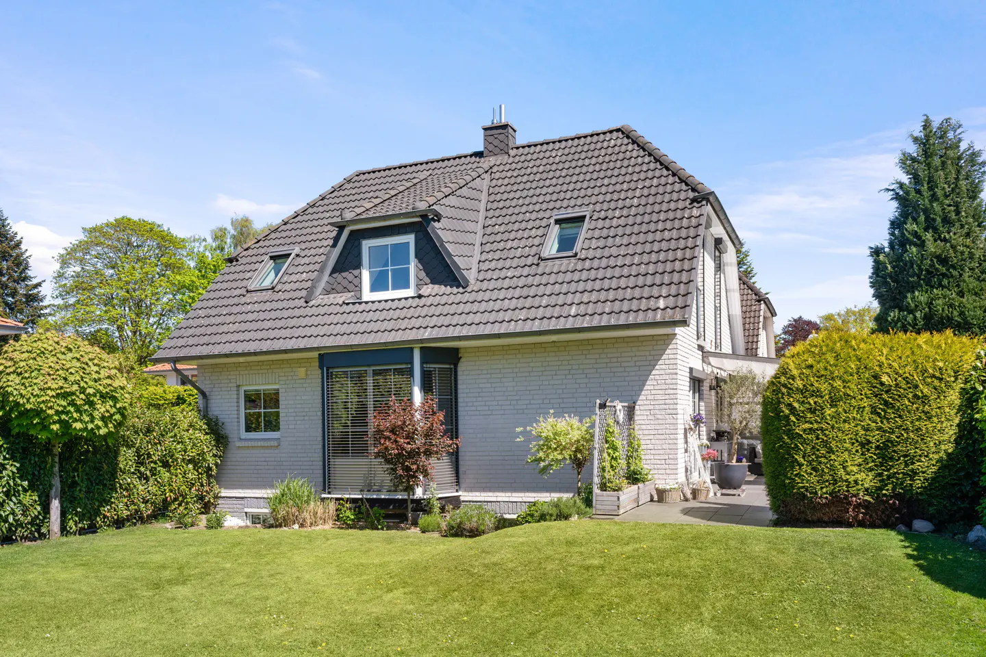 A two-story white brick house with a dark tiled roof, dormer windows, and a lush green lawn under a clear blue sky.