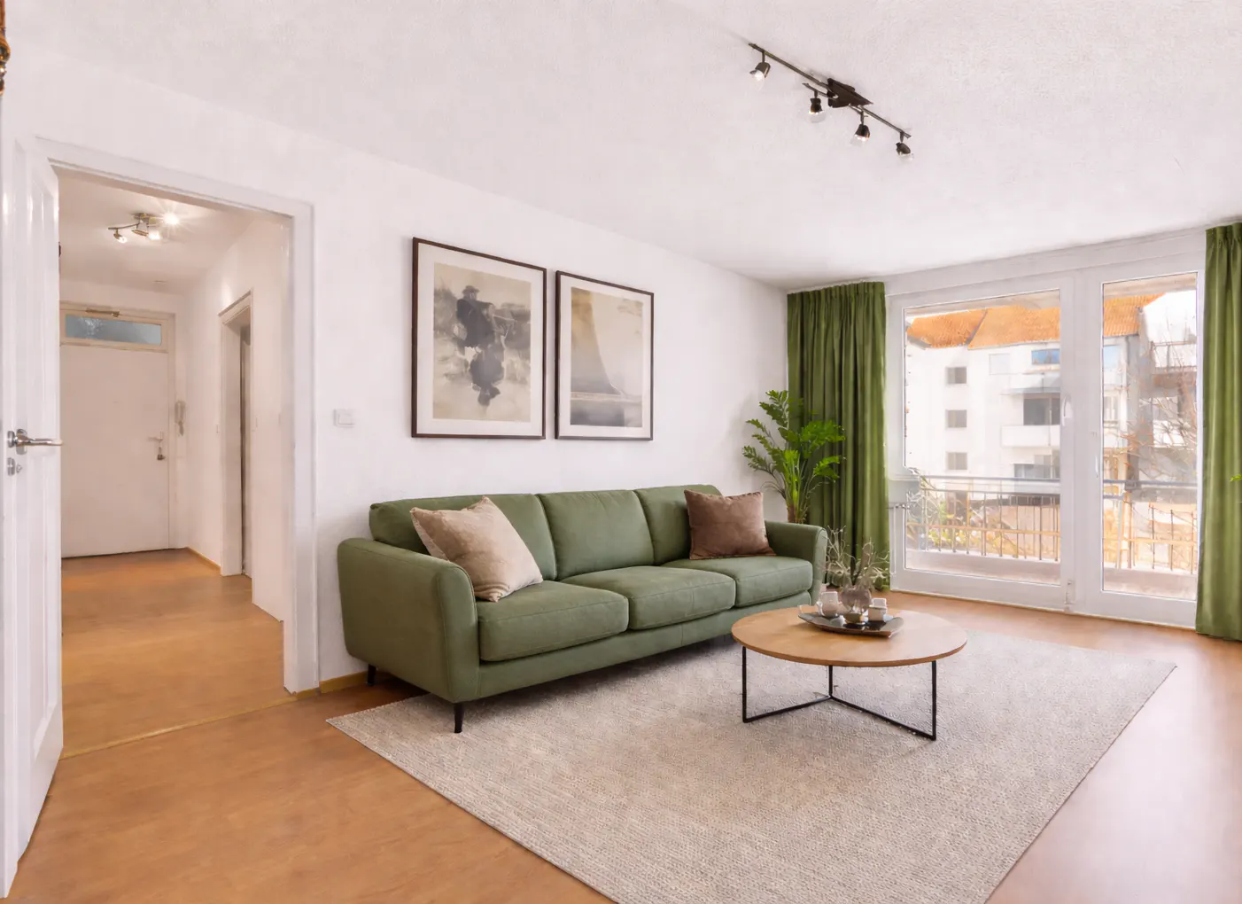 Living room with a green sofa, round wood table, and beige rug. Two framed pictures hang above the sofa. Green curtains frame a window.