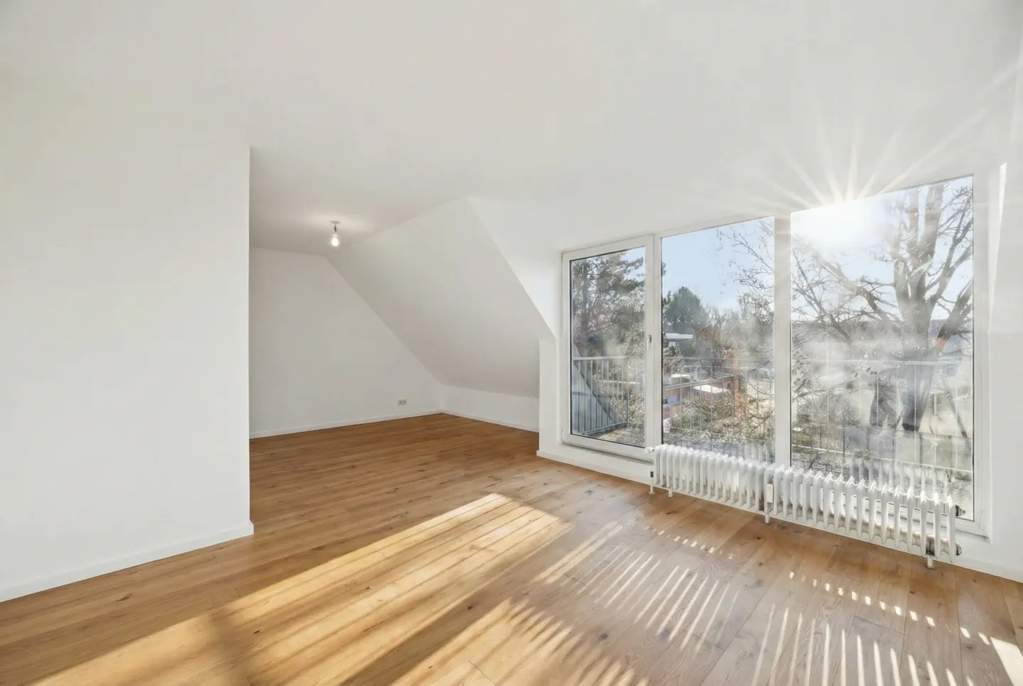 Bright, empty attic room with wood floors, white walls, and a large window with a radiator. Sunlight streams in.