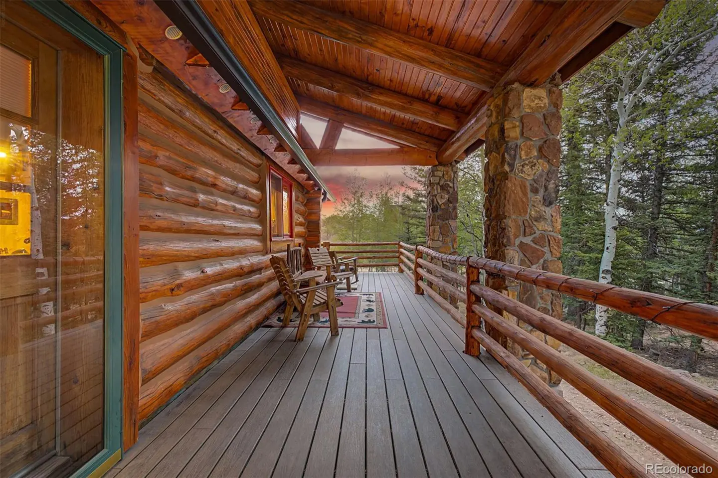 Covered porch of a log cabin with wooden chairs, stone pillars, and a view of trees at sunset.