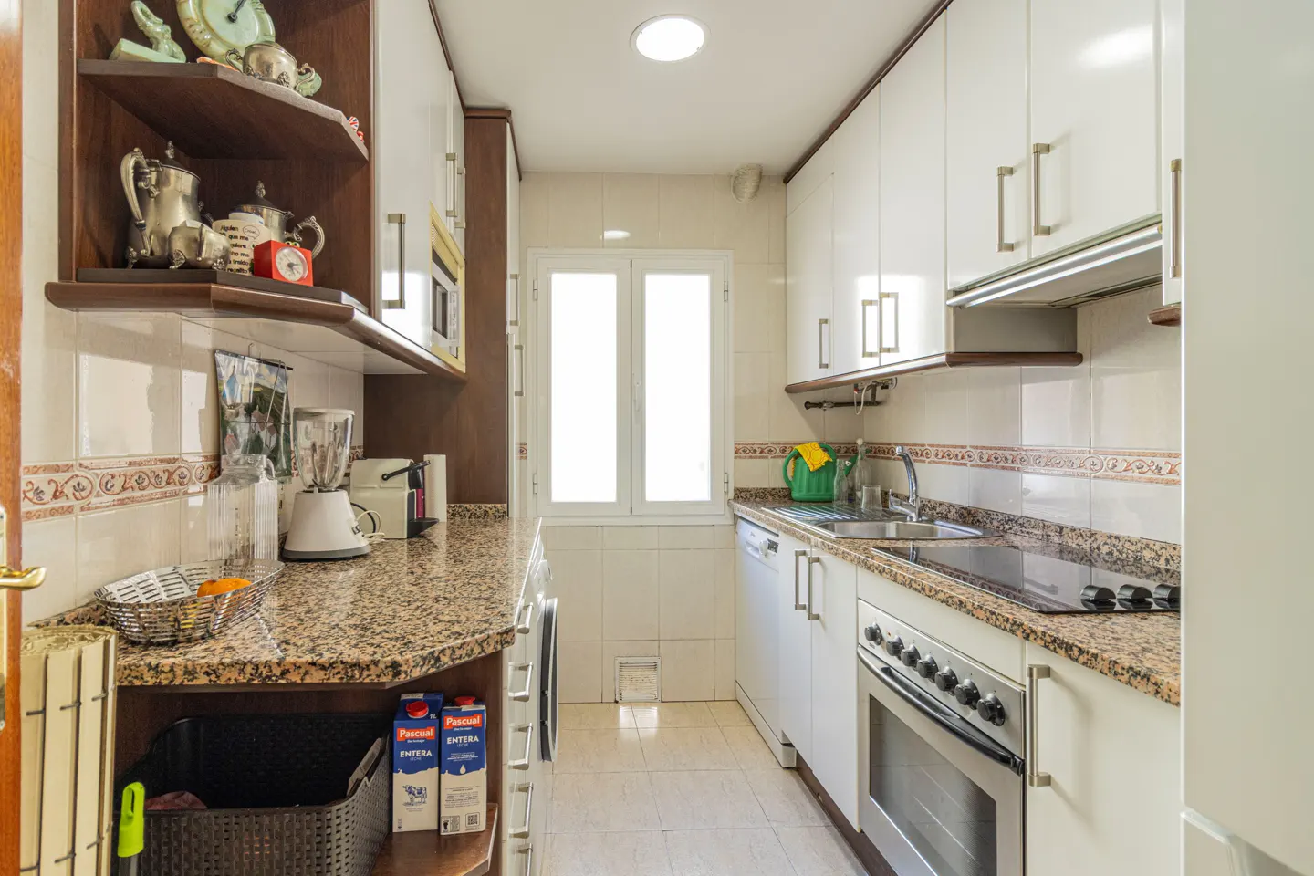 A galley kitchen with white cabinets, granite countertops, and a window. Shelves display decorative items.