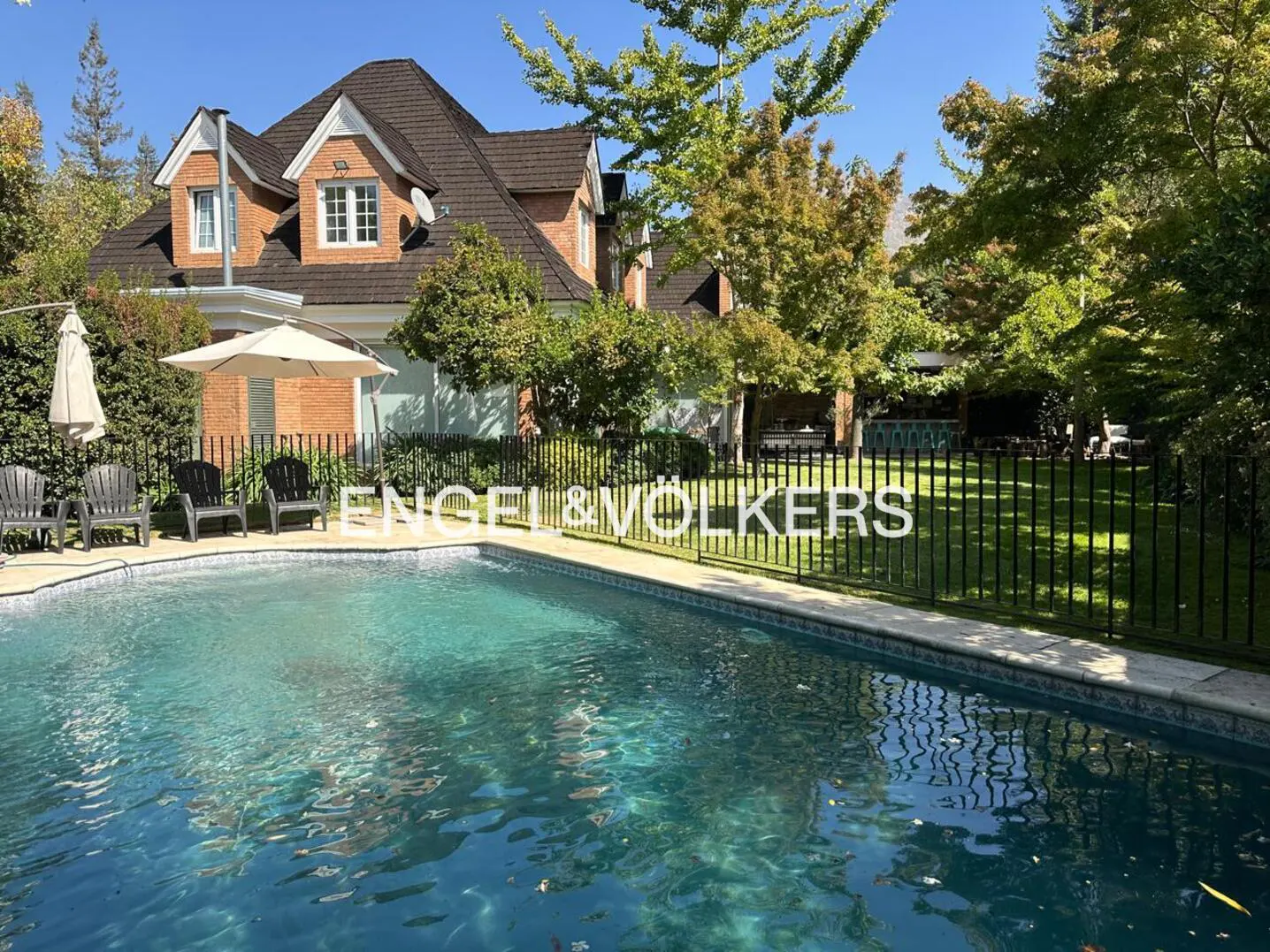 A backyard pool with chairs, umbrellas, and a brick house with a brown roof in the background. Trees surround the property.