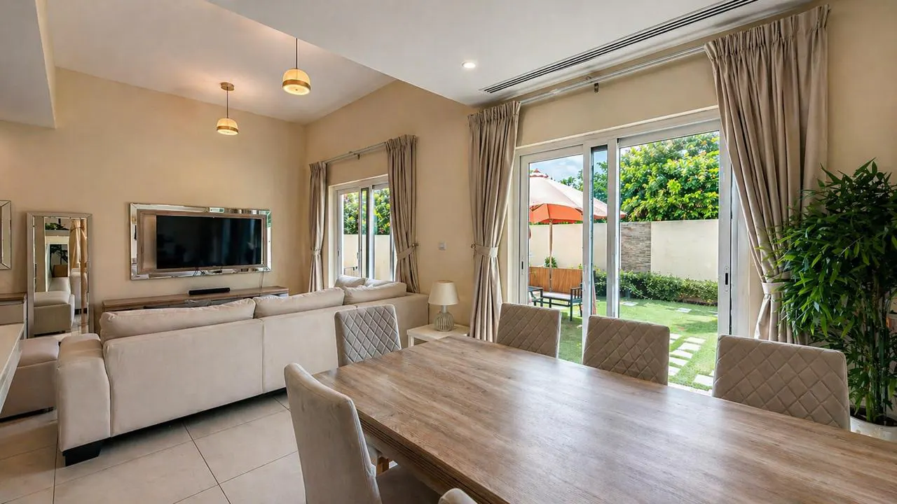 A bright living room with a wood table, beige sofa, and a view of a green backyard with an orange umbrella.
