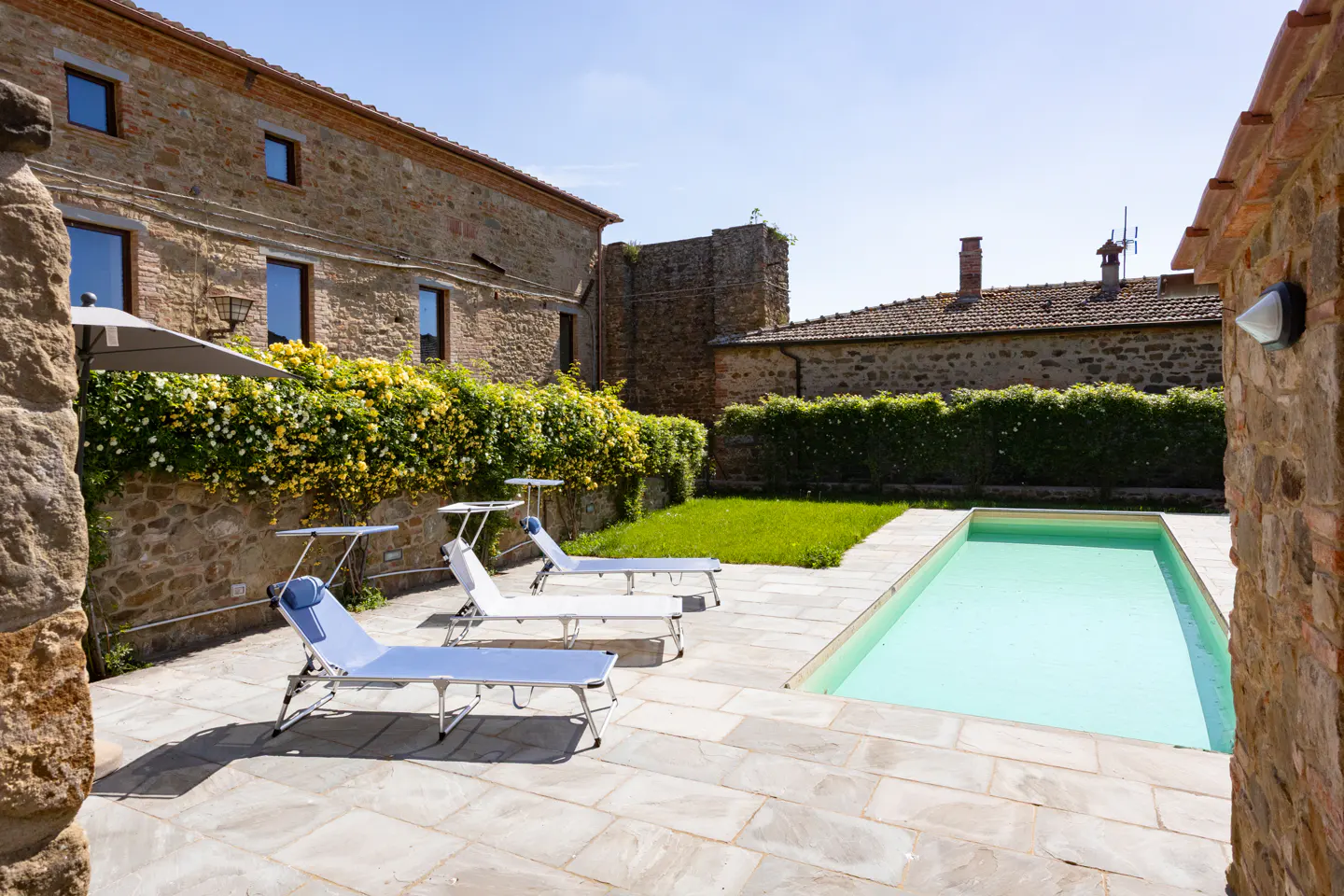 Outdoor pool area with stone buildings, lounge chairs, and green grass. The pool is rectangular and filled with light blue water.