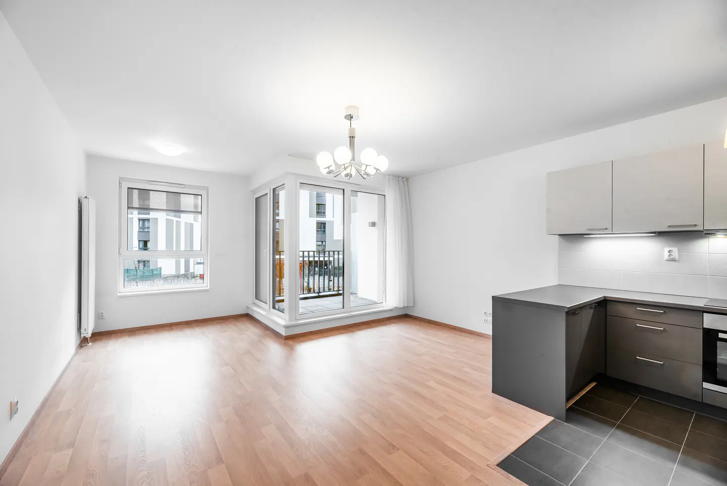 Bright open-plan living area with light wood floors, a modern grey kitchen, and a balcony with large glass doors.