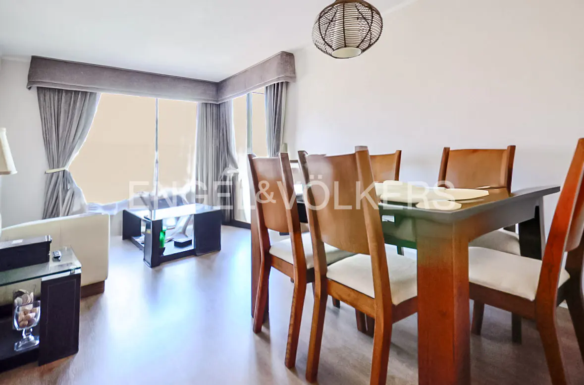 A bright living and dining area with a wooden table, chairs, and a modern light fixture. Gray curtains frame a large window.