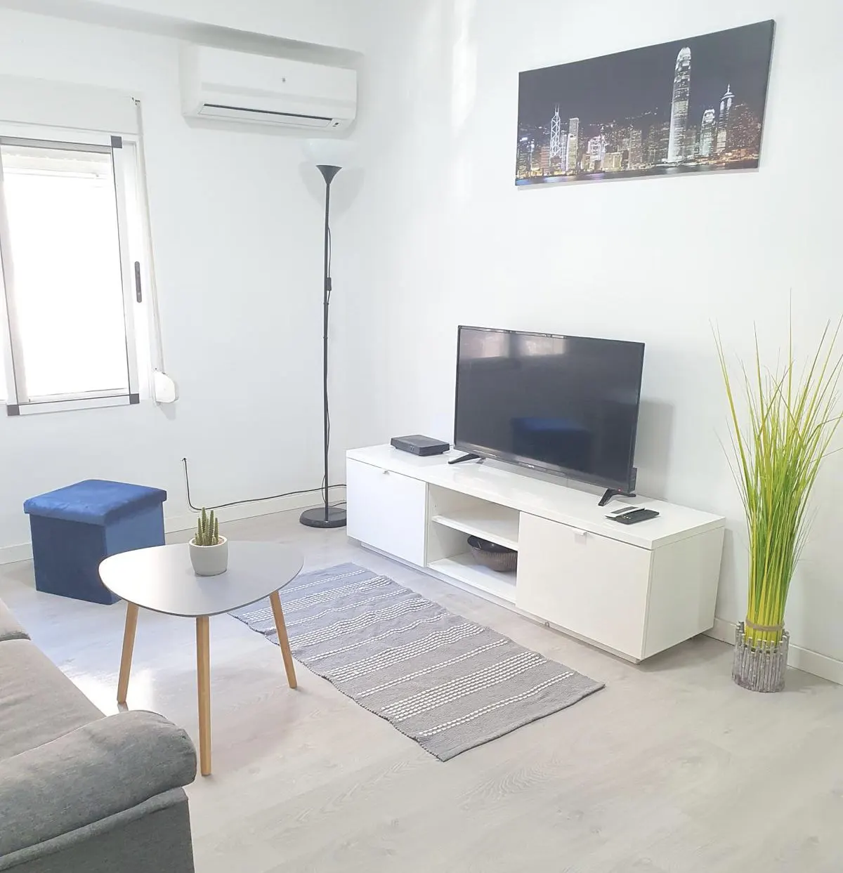 Modern living room with white walls, a TV on a white console, a grey coffee table, a blue ottoman, and a city skyline print.