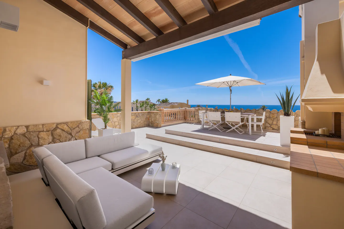 Outdoor patio with white furniture, including a sectional sofa and dining set under an umbrella, overlooking the ocean on a sunny day.