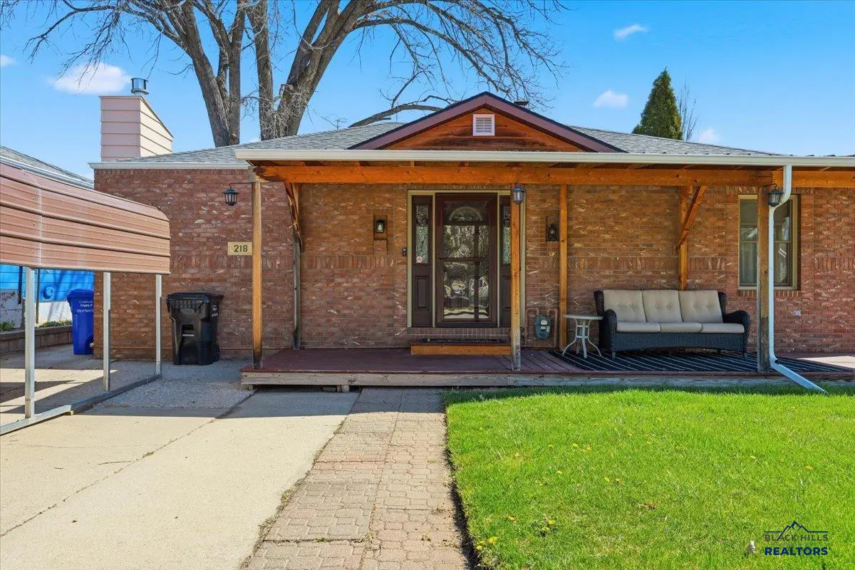 Brick house with a covered porch, a brown door, and a beige sofa. A carport is on the left, and green grass is in the front yard.