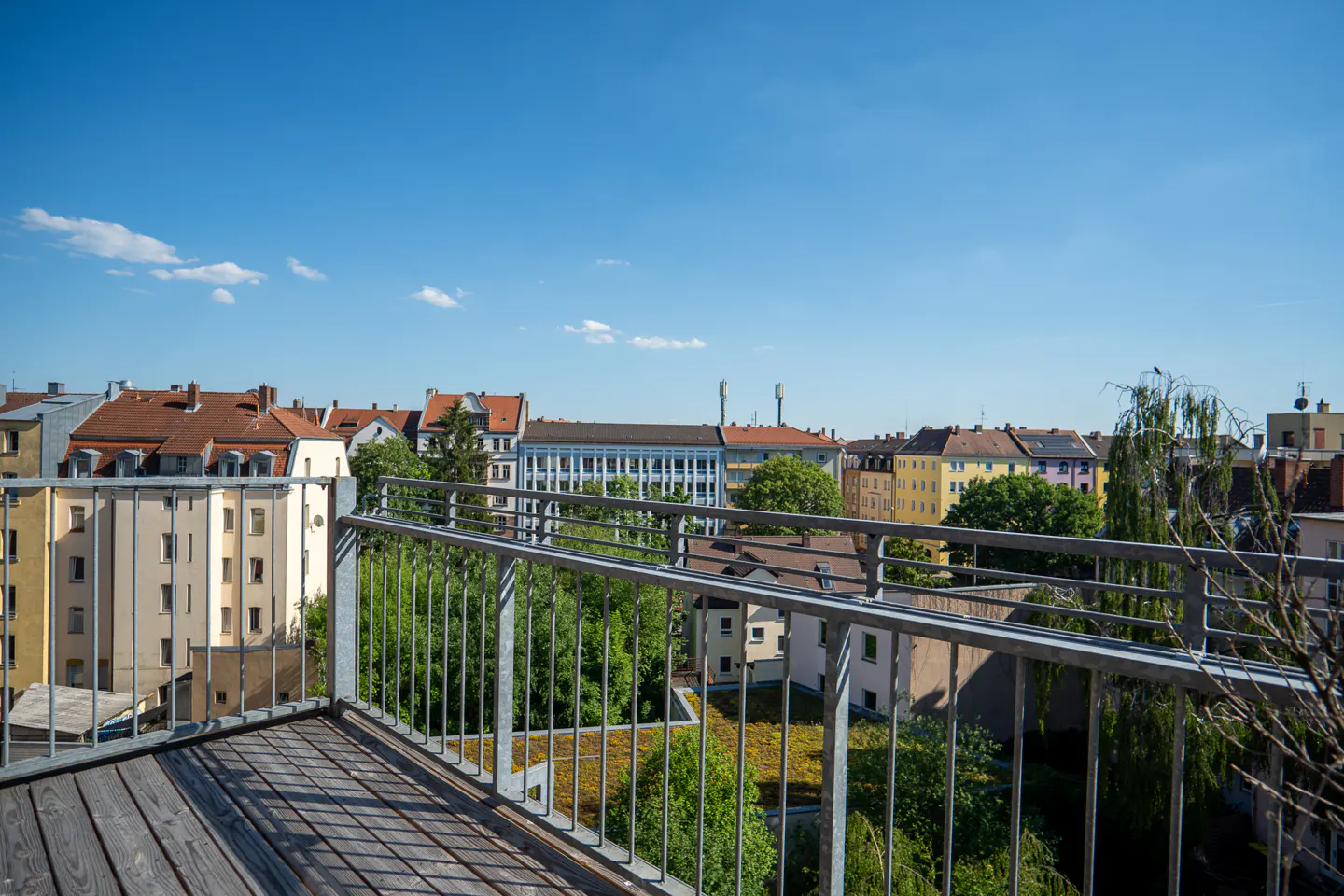 View from a wooden balcony with a metal railing overlooking a European city with colorful buildings and green trees under a blue sky.
