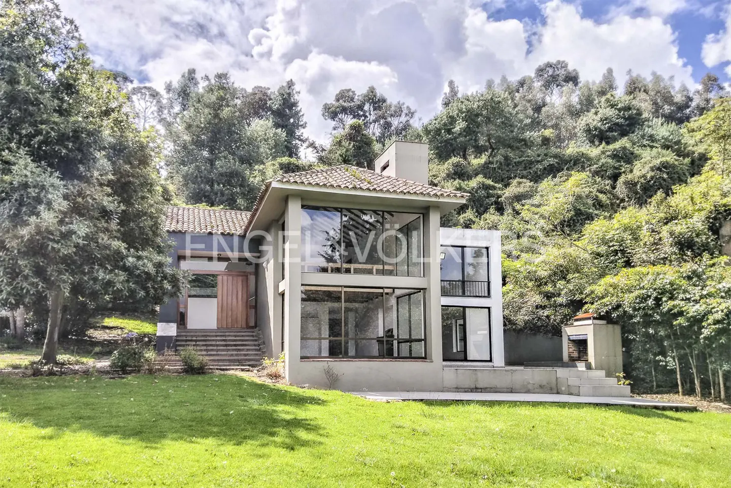 Two-story modern house with large windows, a tile roof, and a green lawn, surrounded by trees and a cloudy sky.