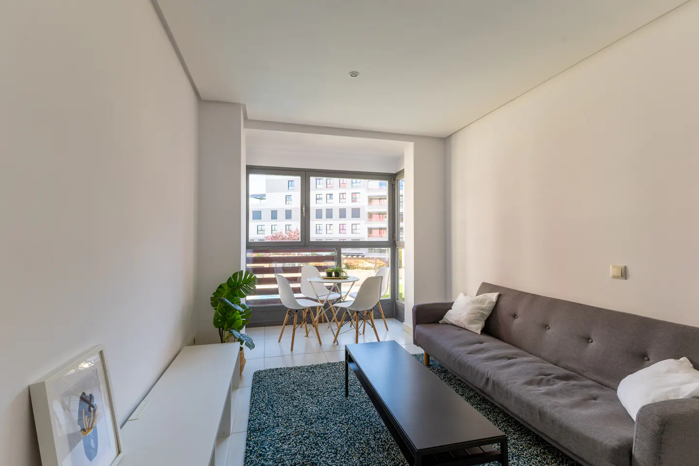 Modern living room with gray sofa, black coffee table, and dining area by a large window. Neutral walls and a blue rug complete the space.