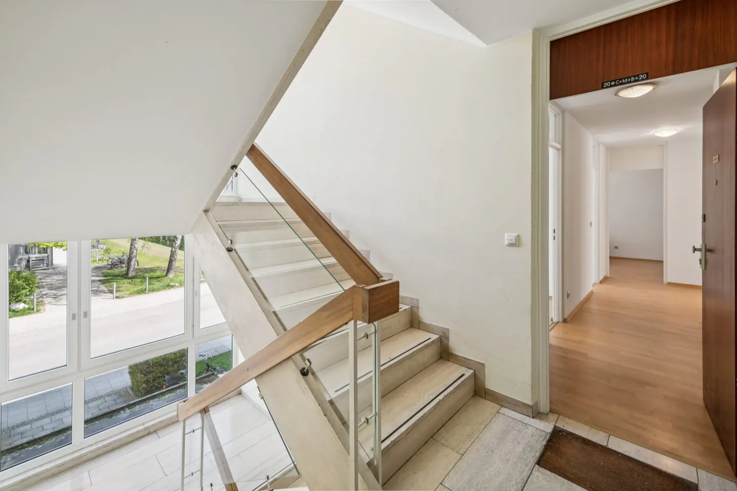 Interior view of a bright stairwell with white walls, light stone stairs, wood and glass railings, and a view to the outside. An open door reveals a hallway with wood floors.