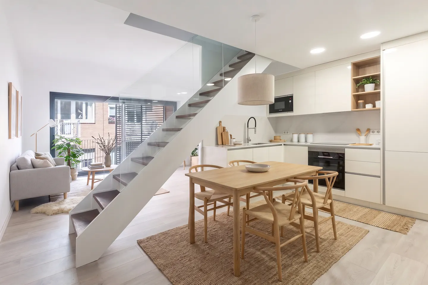 Bright, modern apartment interior with white walls, light wood floors, and open staircase. Dining table, kitchen, and living area visible.
