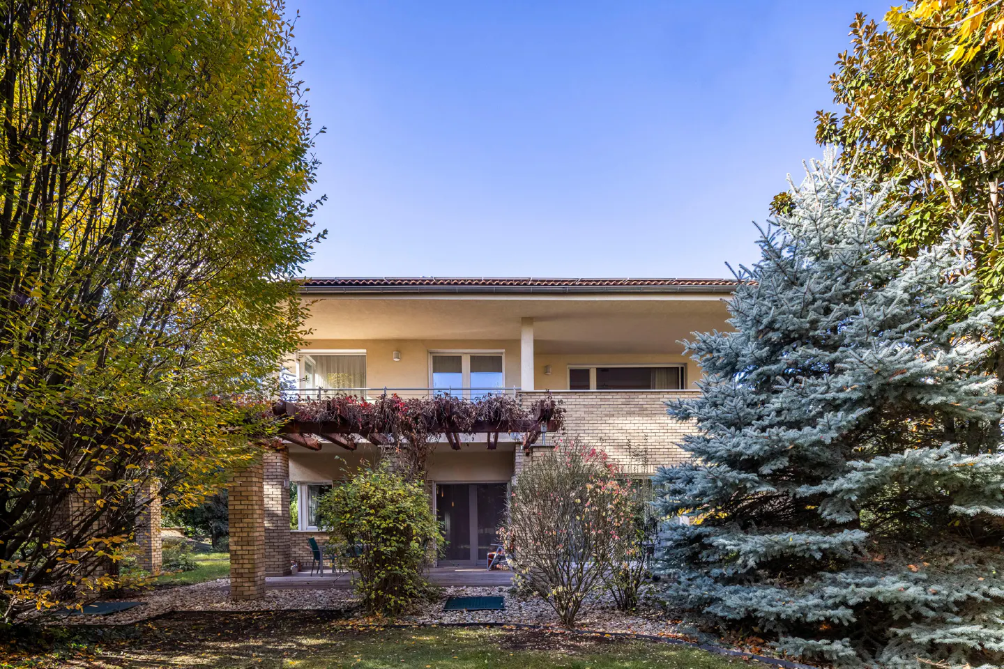 Two-story house with a balcony, surrounded by trees and greenery under a clear blue sky.