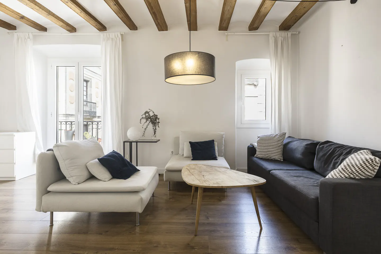 Bright living room with white walls, wood floors, and exposed beams. A gray sofa and white loveseat face a wood coffee table. A black pendant light hangs overhead.