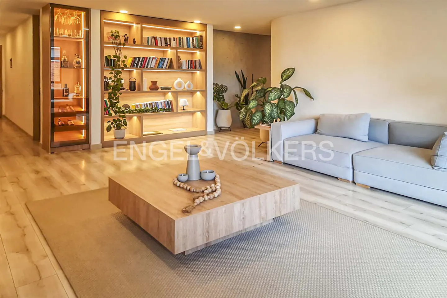 Modern living room with a light wood coffee table, grey sectional sofa, built-in bookshelf, and a lit glass display cabinet.