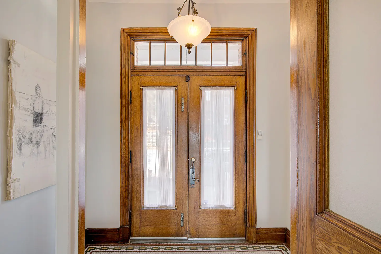 Interior view of a wooden double door with sheer curtains, topped by a transom window and a vintage pendant light.