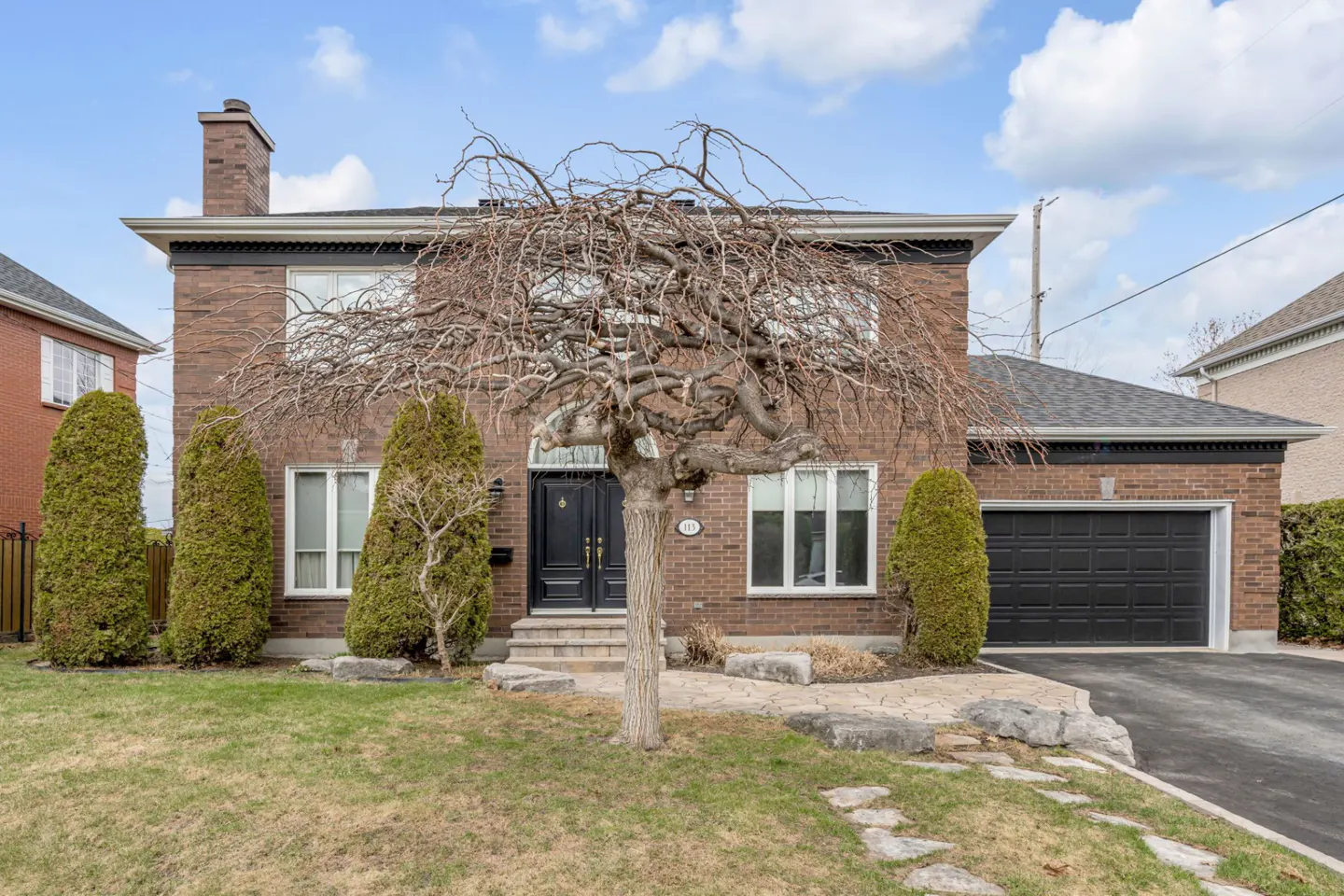 Two-story brick house with a black garage door, a bare tree, and green bushes in front.