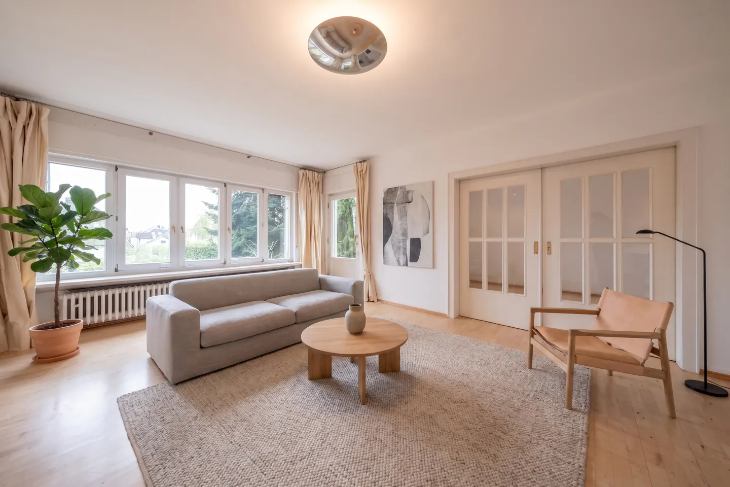 Bright living room with a beige sofa, round wood table, and tan leather chair on a woven rug. Large windows and sliding glass doors let in natural light.