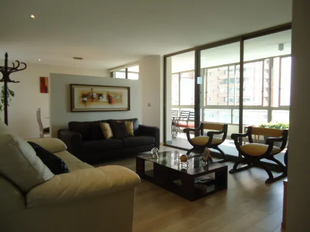 A bright living room with a black sofa, cream sofa, dark wood chairs, and a glass coffee table. A balcony is visible through large windows.