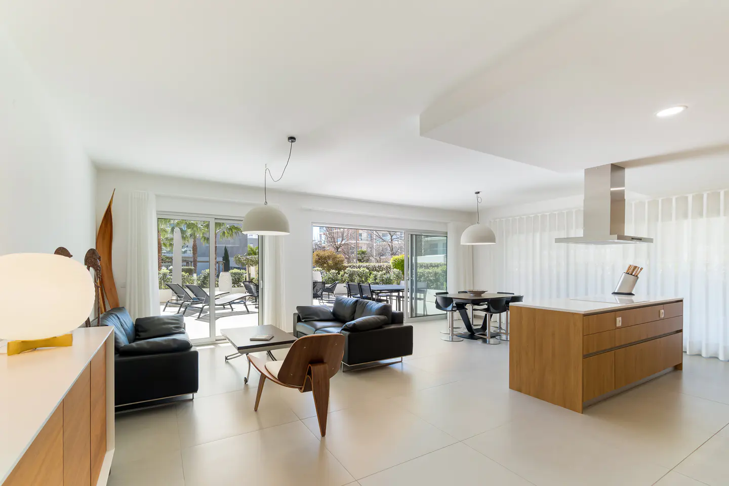 Bright, open-plan living space with black sofas, wood chair, kitchen island, and outdoor patio visible through sliding glass doors.