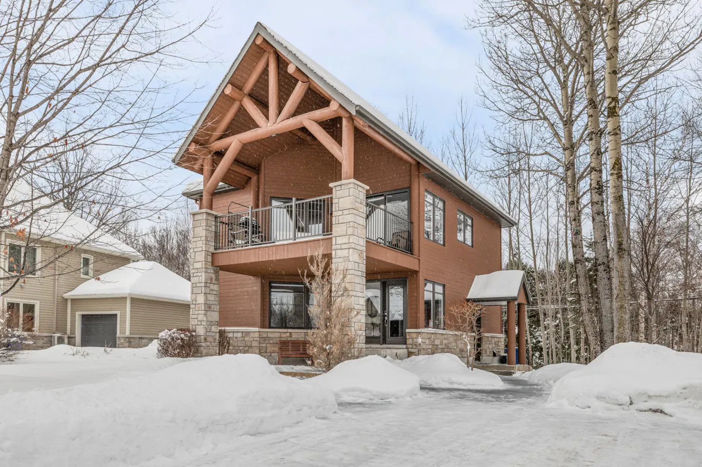 Two-story brown house with a balcony and log beams, surrounded by snow-covered ground and bare trees.