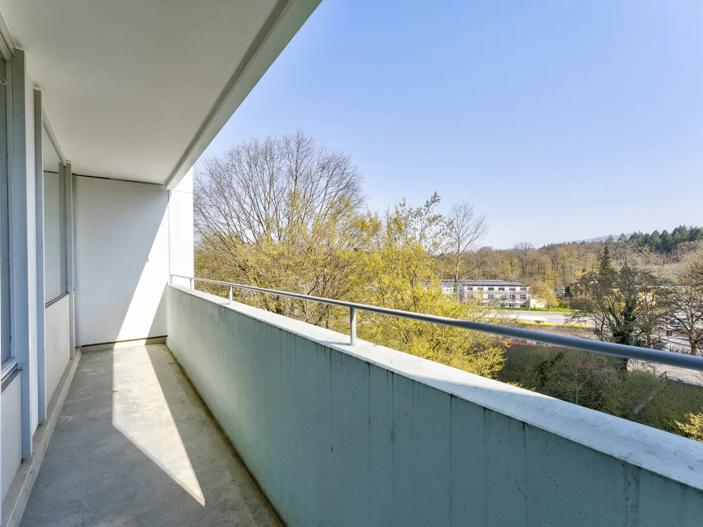 Balcony view with a concrete floor and railing overlooking trees and a building on a sunny day.