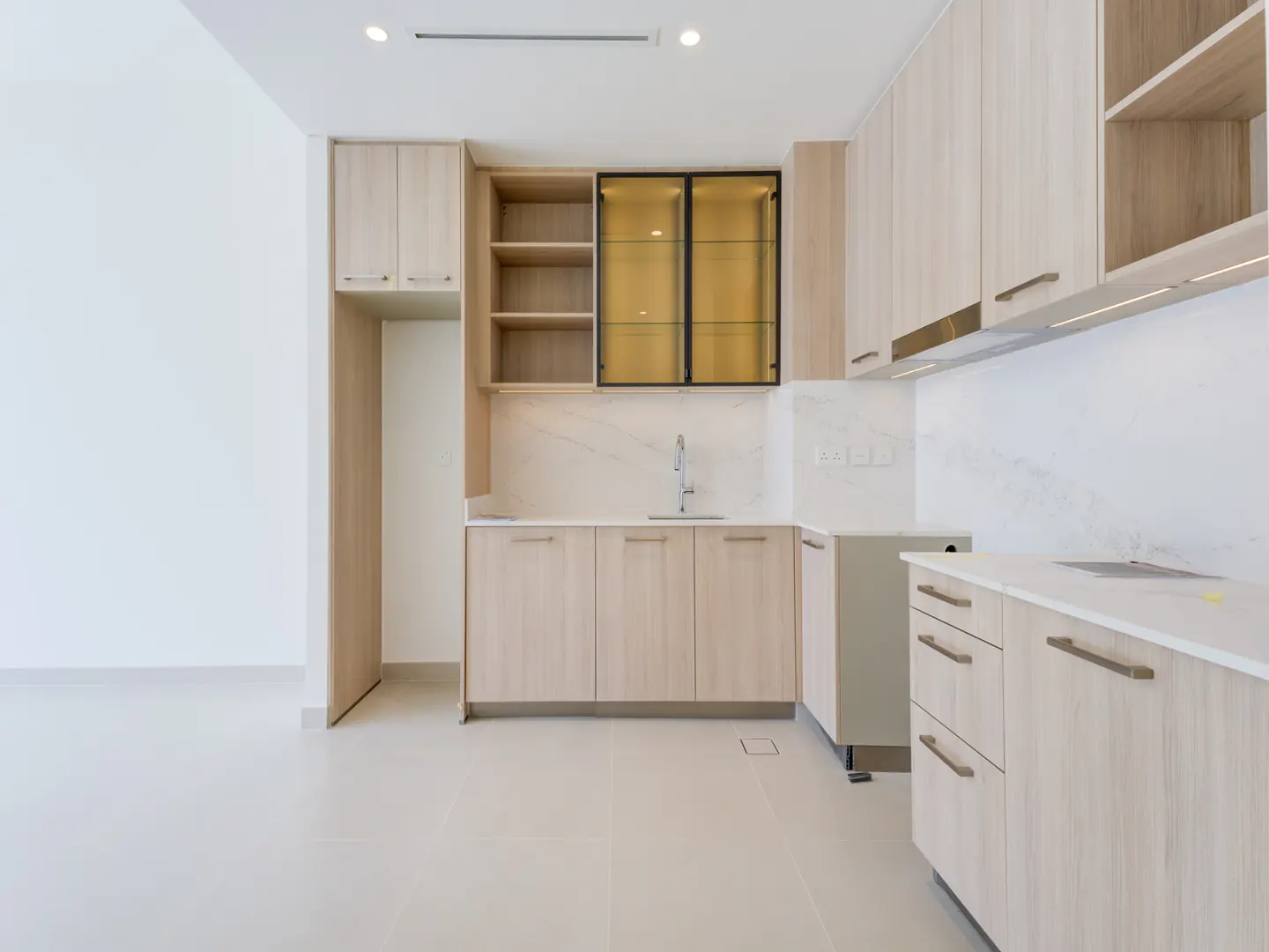 A bright kitchen with light wood cabinets, white countertops, and stainless steel sink. Open shelves and a glass-front cabinet add visual interest.