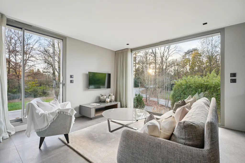 Living room with gray sofa, chair, and TV. Large windows overlook trees and greenery. Neutral tones create a modern, bright space.