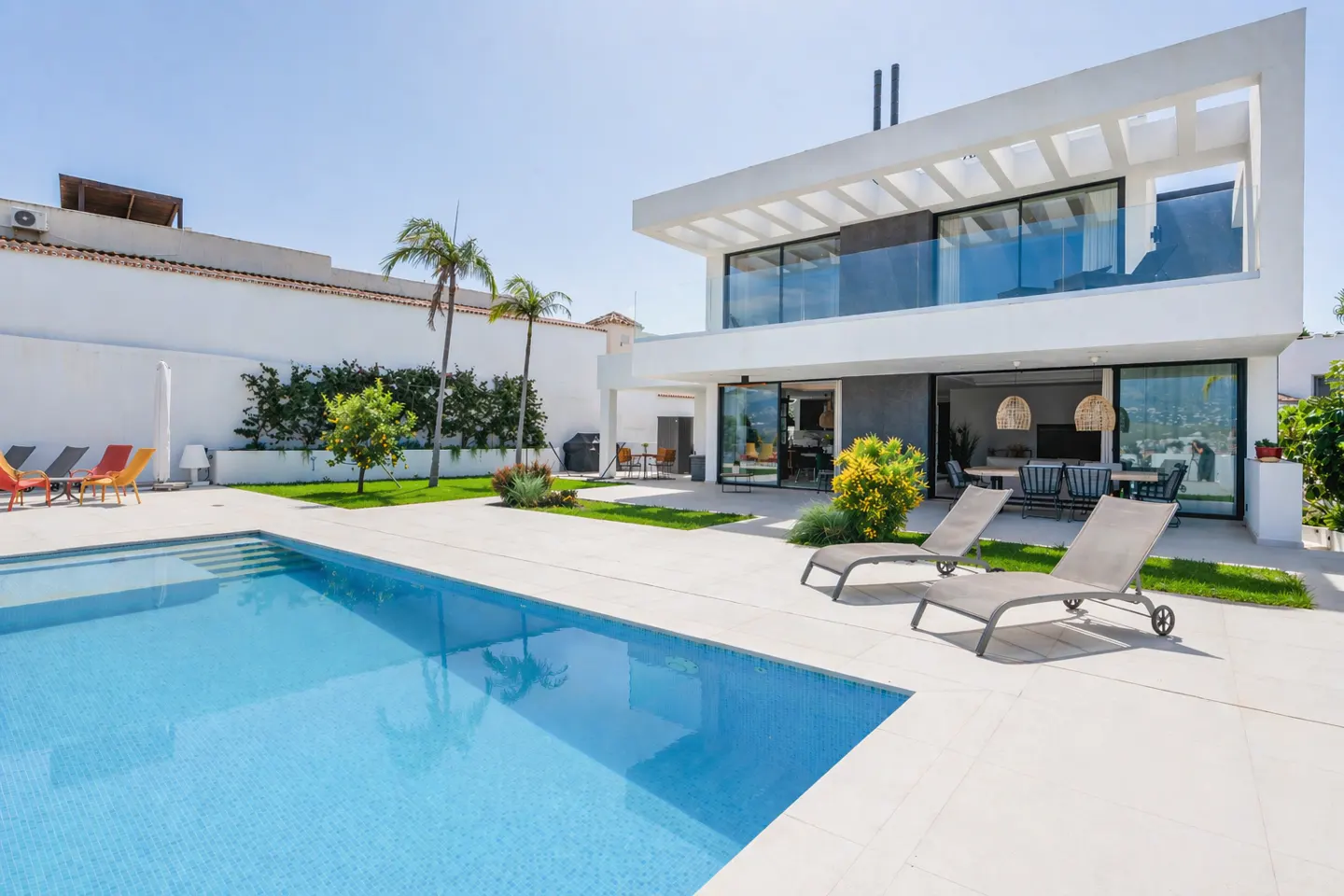 Modern white house with a blue pool, two lounge chairs, and palm trees under a clear blue sky.