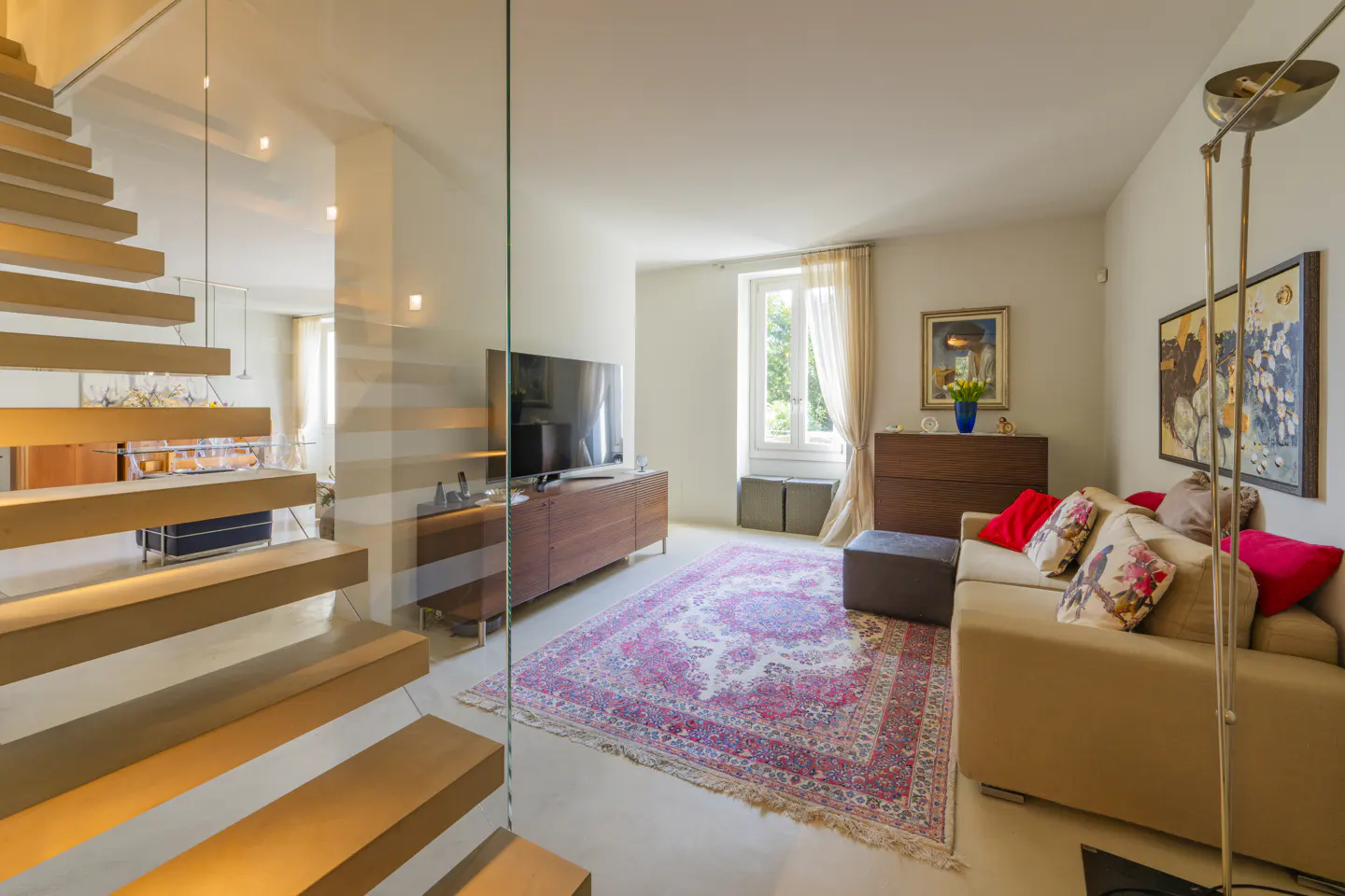 Bright living room with a beige sofa, colorful rug, and wooden stairs with glass railing. Paintings adorn the walls.