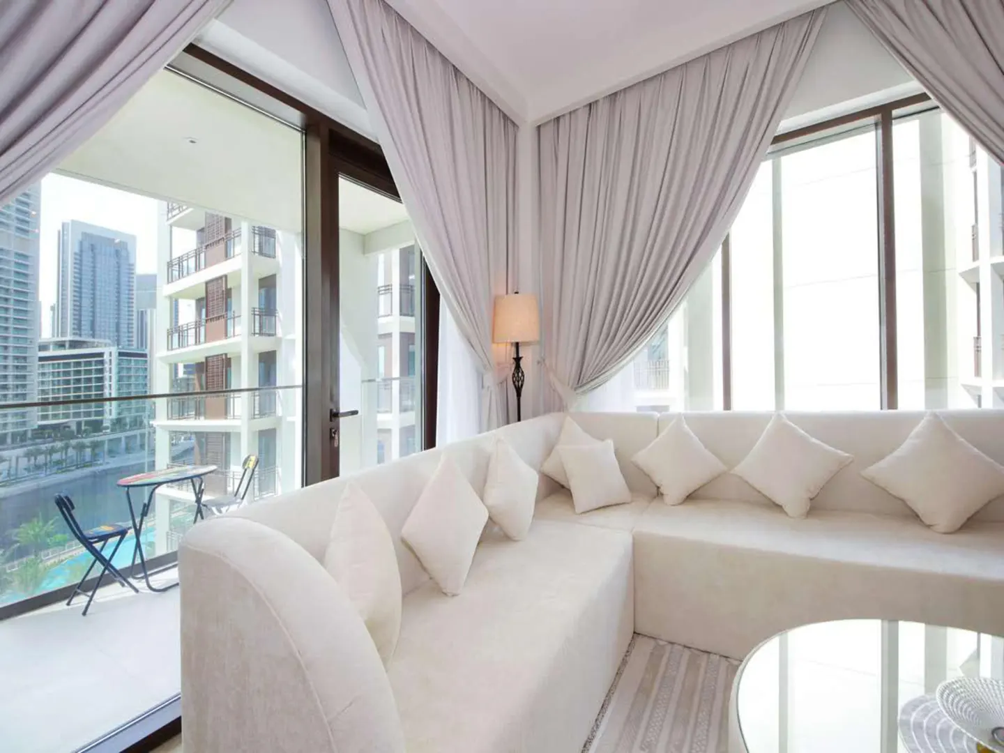 Bright living room with white L-shaped sofa, square pillows, and glass table. Balcony view of city buildings and water. Gray curtains frame the windows.