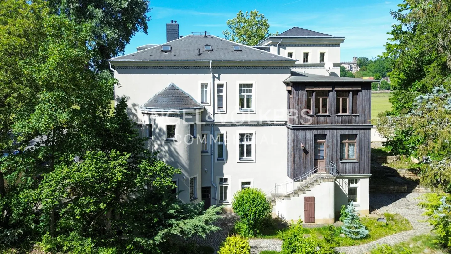 Exterior view of a light gray building with a dark gray roof and a dark wood addition, surrounded by green trees.