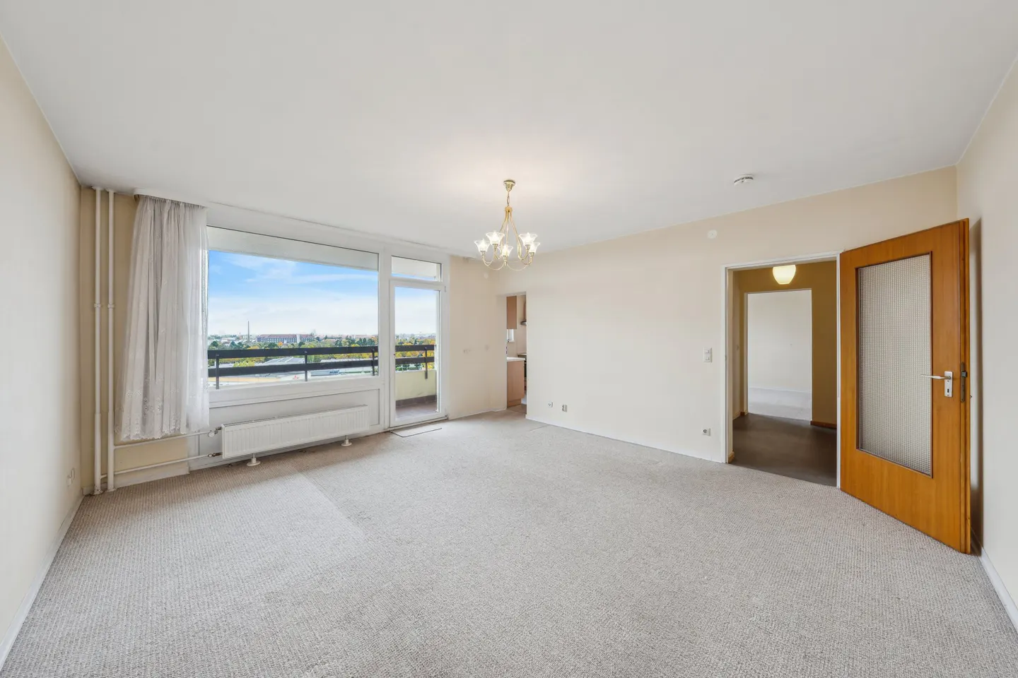 Bright, empty living room with gray carpet, large window to a balcony, and a chandelier. A doorway leads to another room.