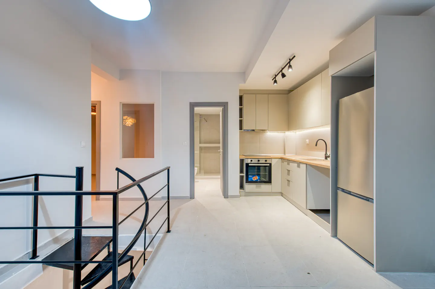 Modern apartment interior with a black spiral staircase, a light beige kitchen with stainless steel appliances, and white walls.