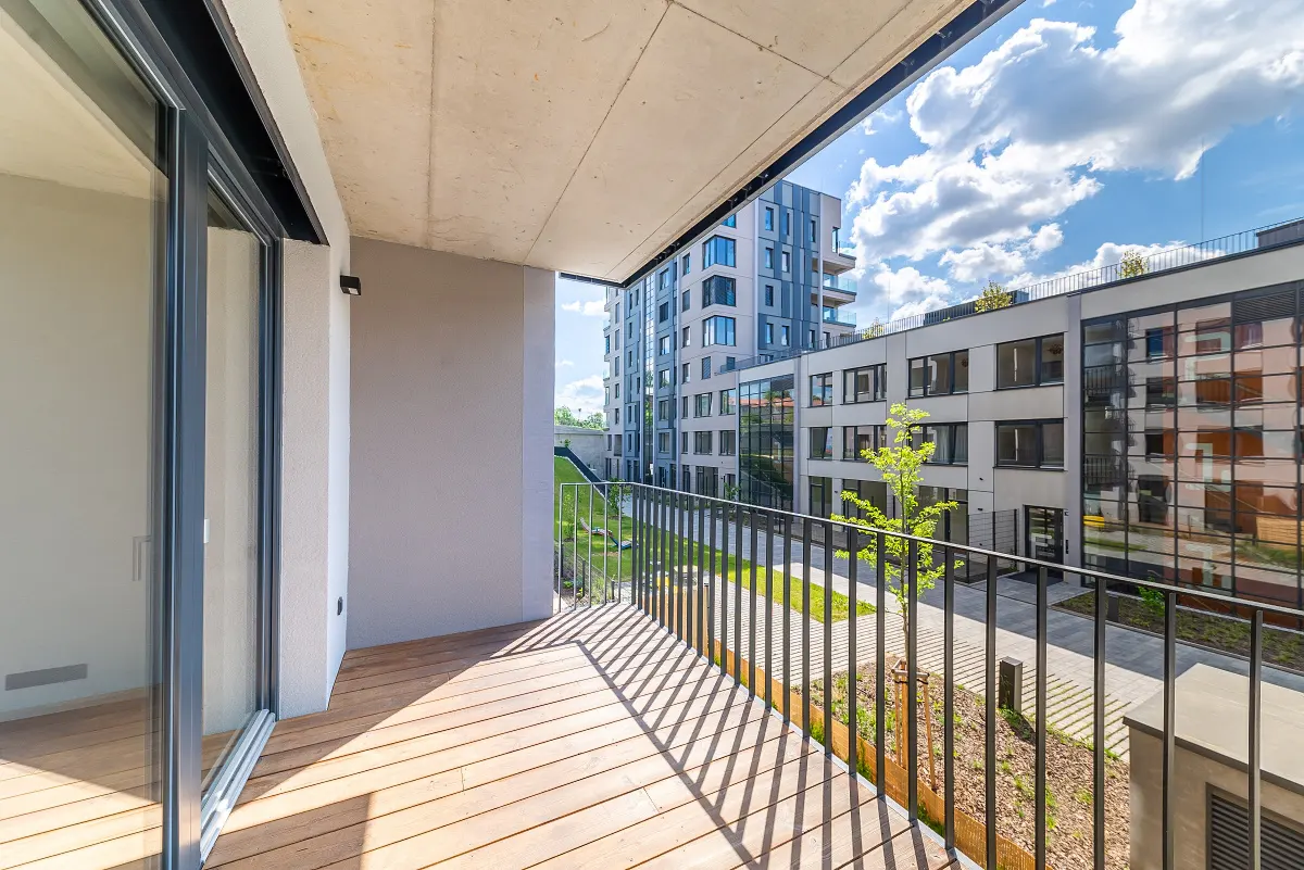 Balcony view with wood floor, black railing, and sliding glass door. Modern buildings and green space visible in the background.
