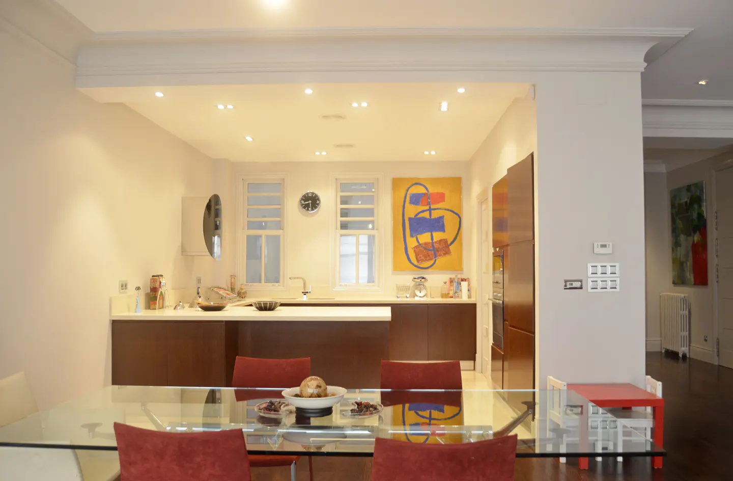 A bright, modern kitchen with a glass dining table and red chairs. An abstract painting hangs on the wall.
