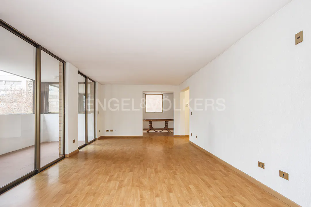 Bright, empty room with wood floors, white walls, and large sliding glass doors to a balcony. A table is visible in the next room.