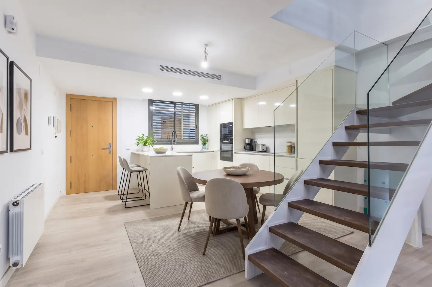 Bright, modern interior with a kitchen, dining area, and staircase. The kitchen has white cabinets and a breakfast bar. A round wooden table with chairs sits nearby. The staircase has glass railings.