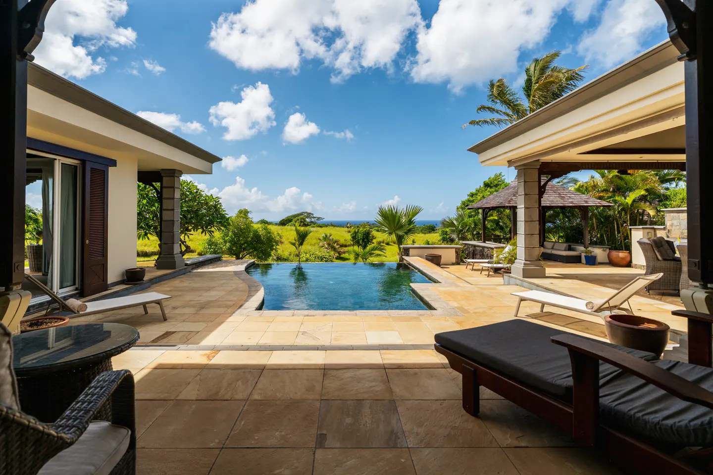Luxury villa patio with pool overlooking green fields and ocean under a blue sky with white clouds. Lounge chairs and outdoor furniture.